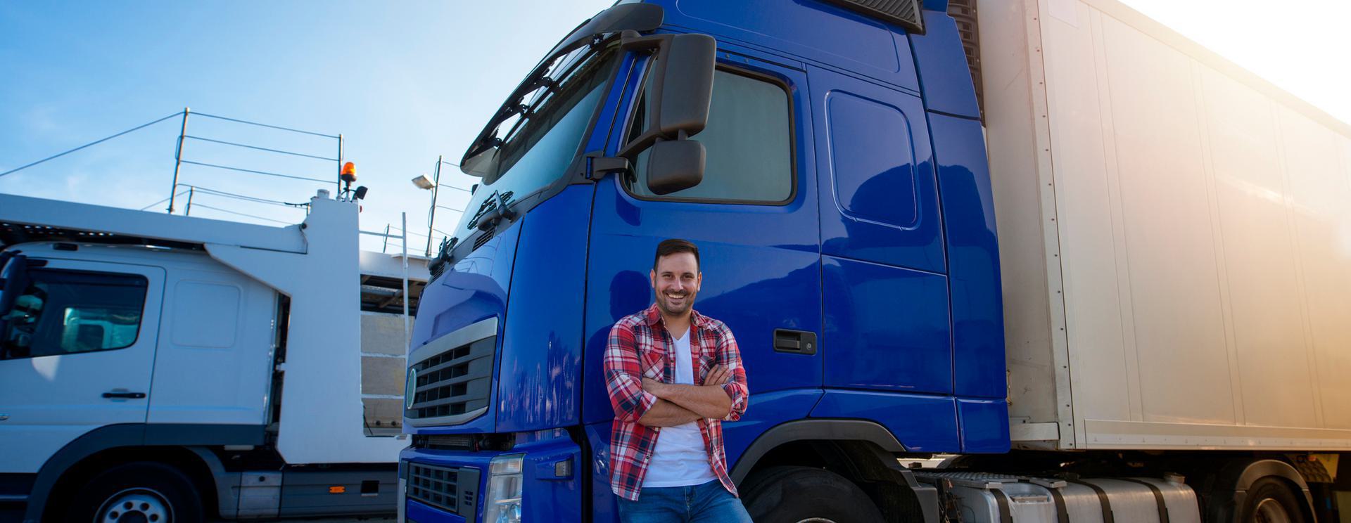 Smiling man in plaid shirt standing beside a blue semi-truck under clear sky.