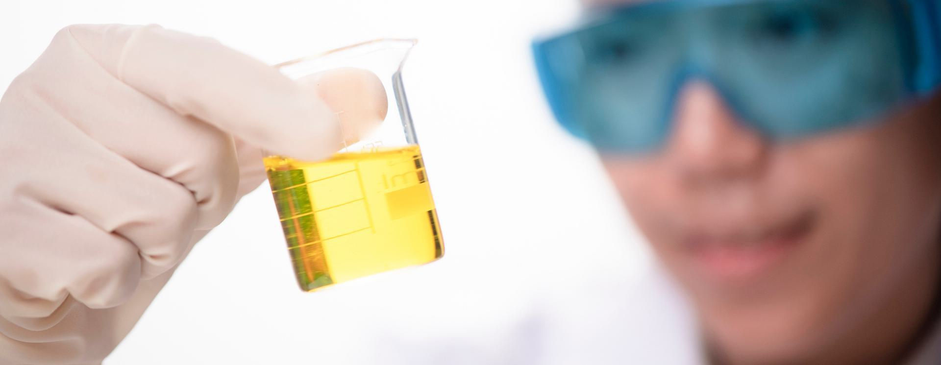 Scientist wearing blue safety goggles examining a yellow liquid in a beaker.
