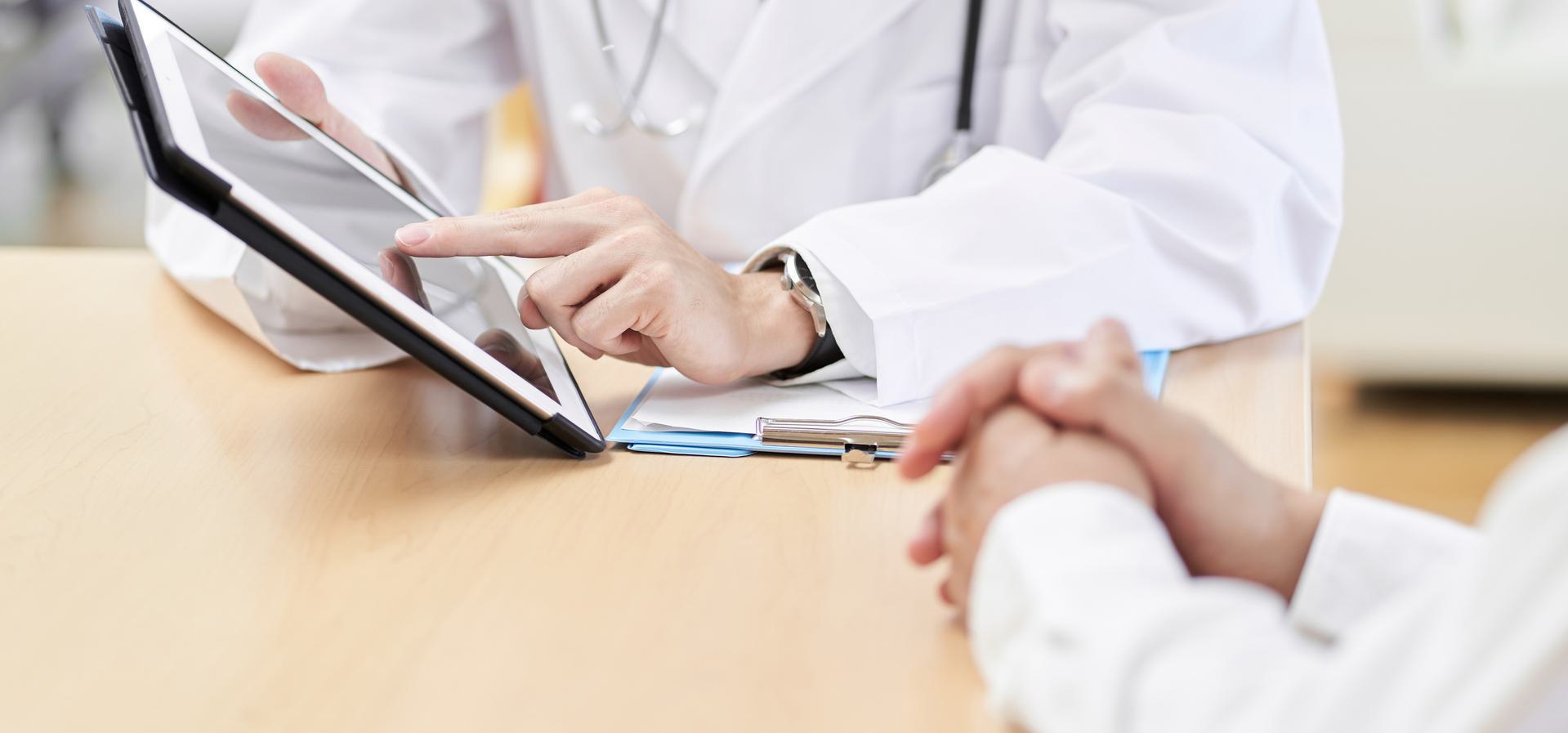 Doctor using a tablet to discuss medical information with a patient at a desk.