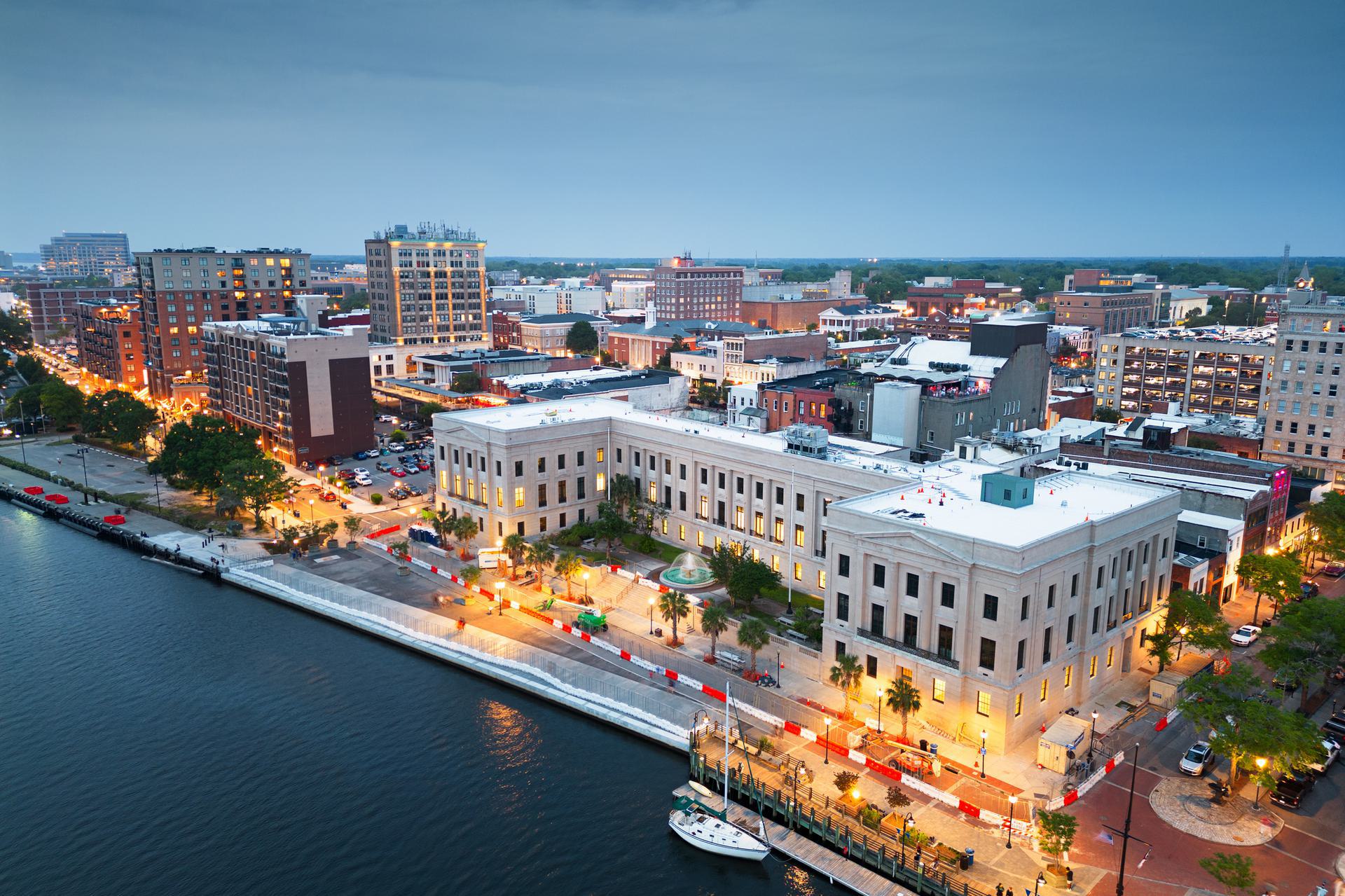 Aerial view of a cityscape at dusk with illuminated buildings along a waterfront.