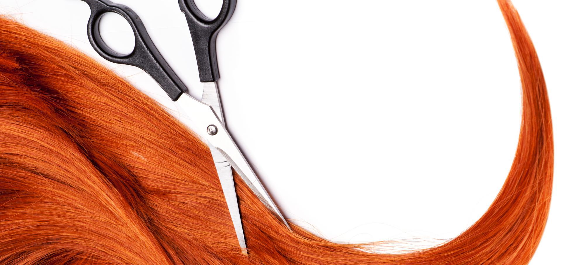 Scissors cutting a strand of vibrant red hair against a white background.