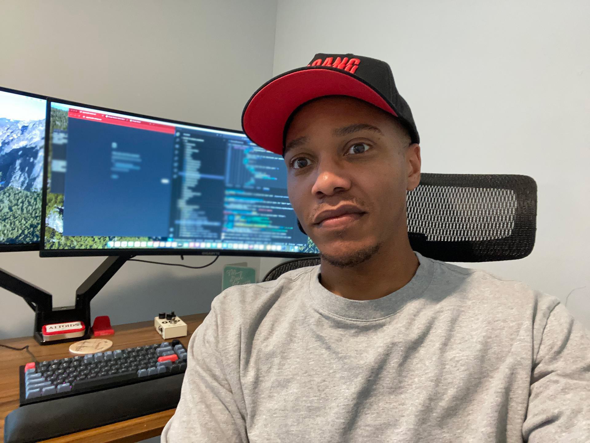 Najee sitting at a desk with dual monitors displaying code, wearing a red and black cap and gray sweatshirt.
