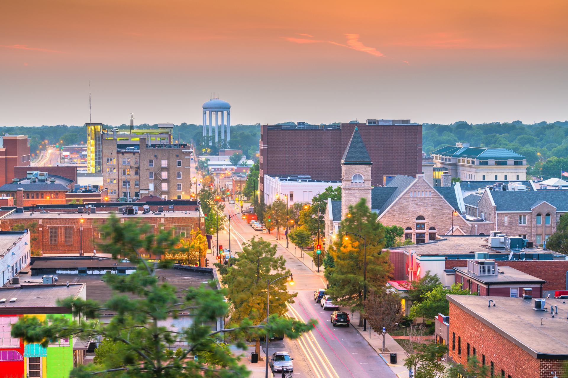 Aerial view of a cityscape at sunset with buildings, streets, and a water tower in the distance.