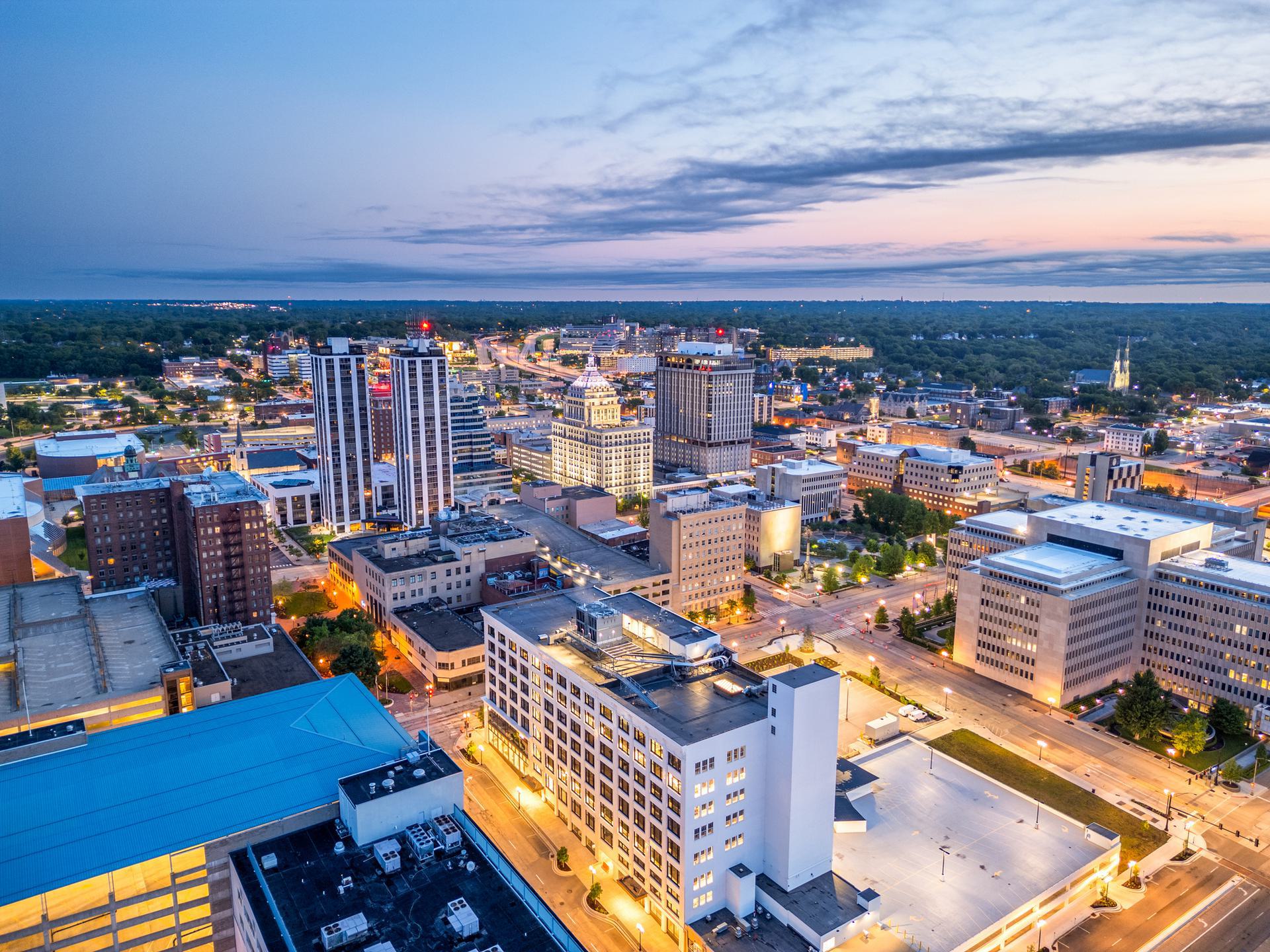 Aerial view of a cityscape at dusk with illuminated buildings and streets, surrounded by greenery under a cloudy sky.