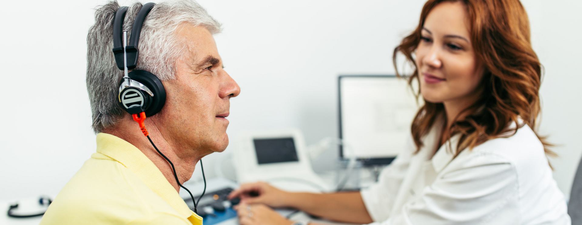Older man wearing headphones during a hearing test with a female audiologist in an office setting.