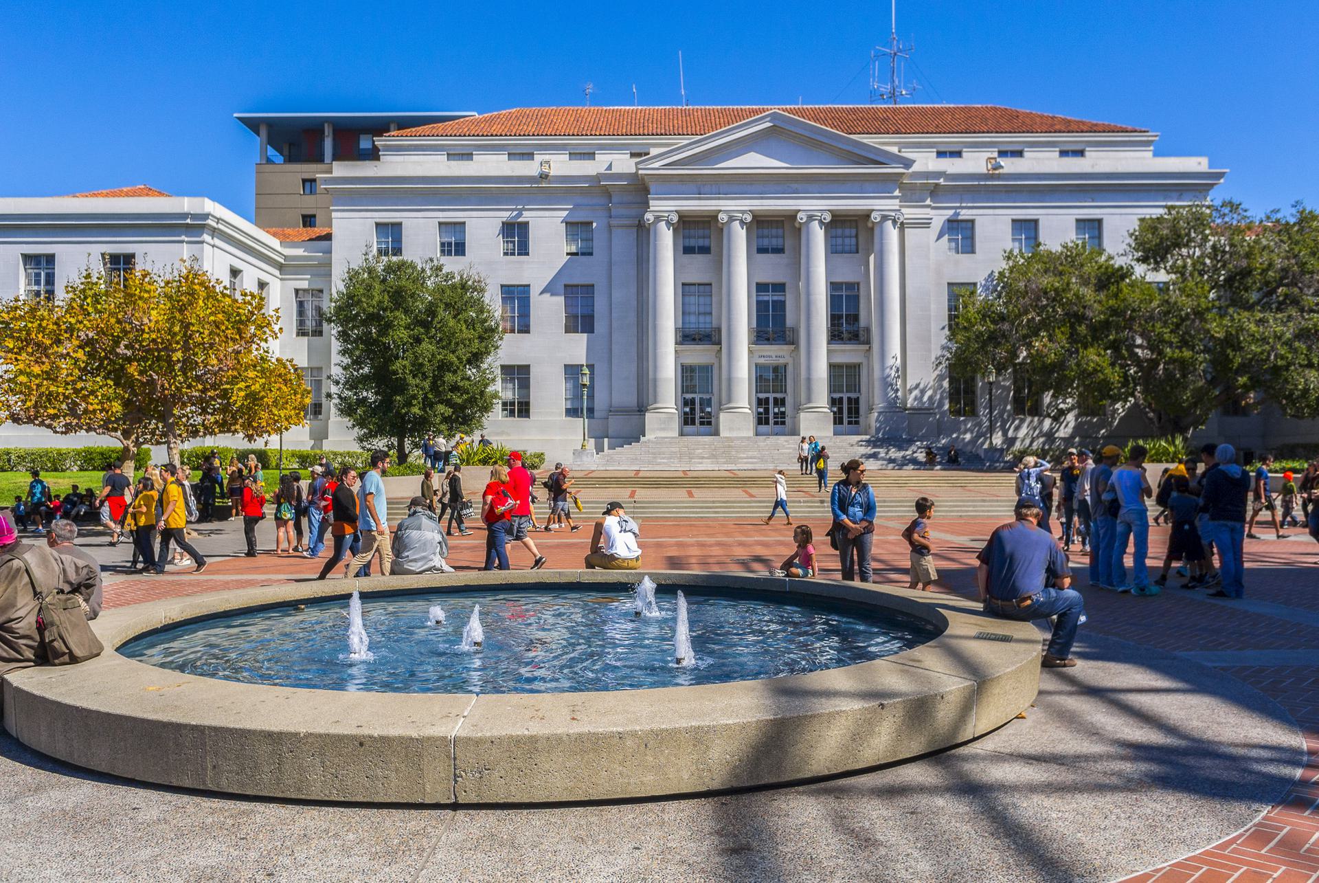 Campus Gathering People gathering around a fountain in front of a large neoclassical building with columns on a sunny day.