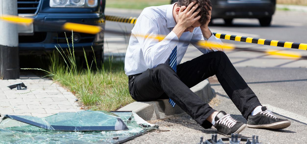 Post Accident Testing Man in distress sitting on curb near car accident scene with broken glass and caution tape.