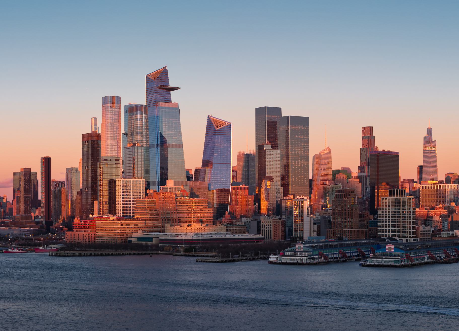 NYC Skyline Sunset New York City skyline at sunset with modern skyscrapers reflecting warm light, viewed from across the Hudson River.