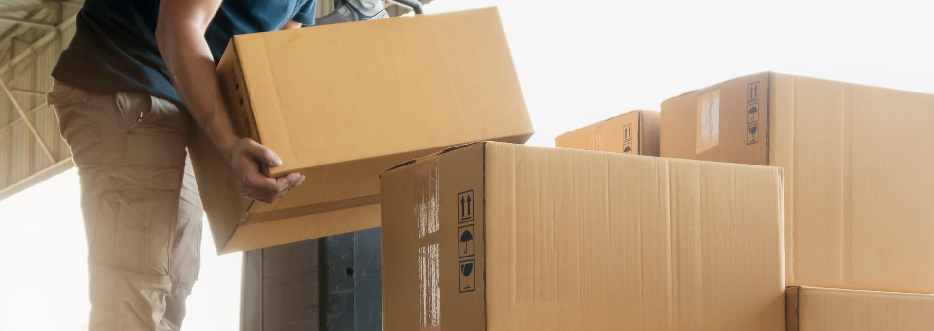 Person lifting a cardboard box in a warehouse with other boxes stacked nearby.