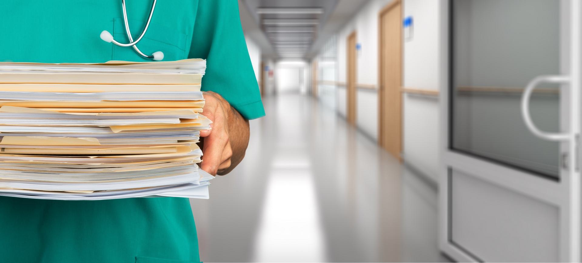 Healthcare professional in scrubs holding a large stack of patient files in a hospital corridor.
