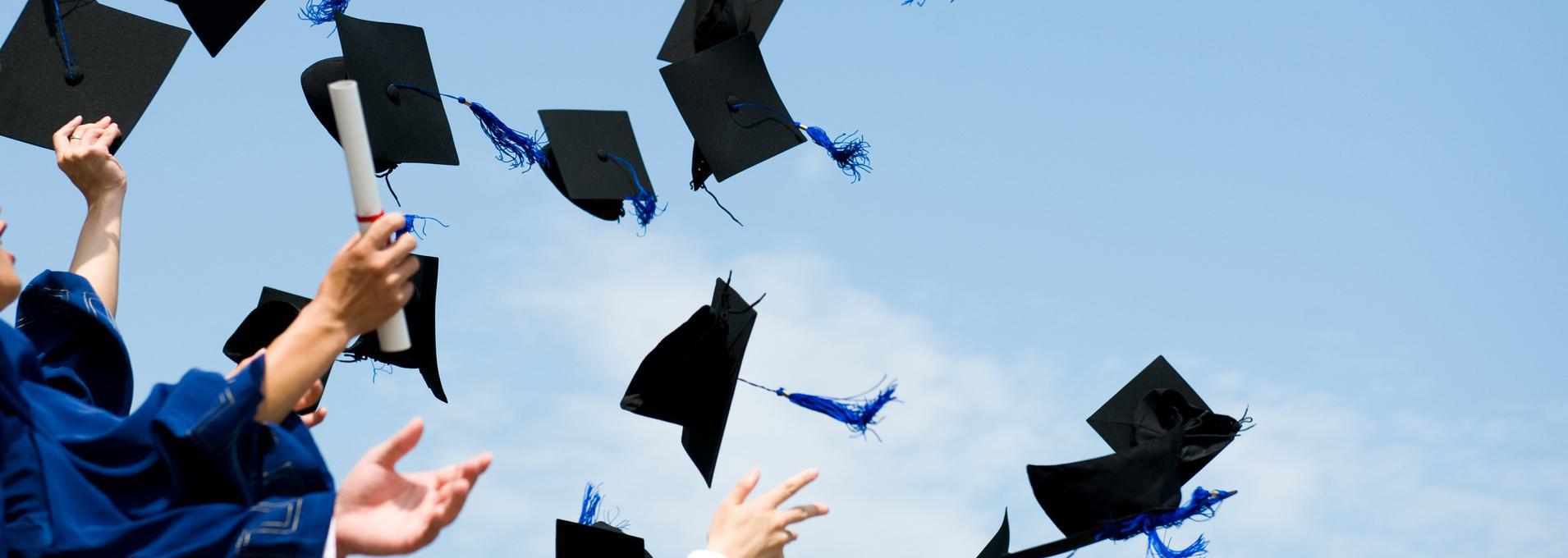 Education History & Degree Verification Graduation caps being thrown in the air by students wearing blue gowns against a clear sky.