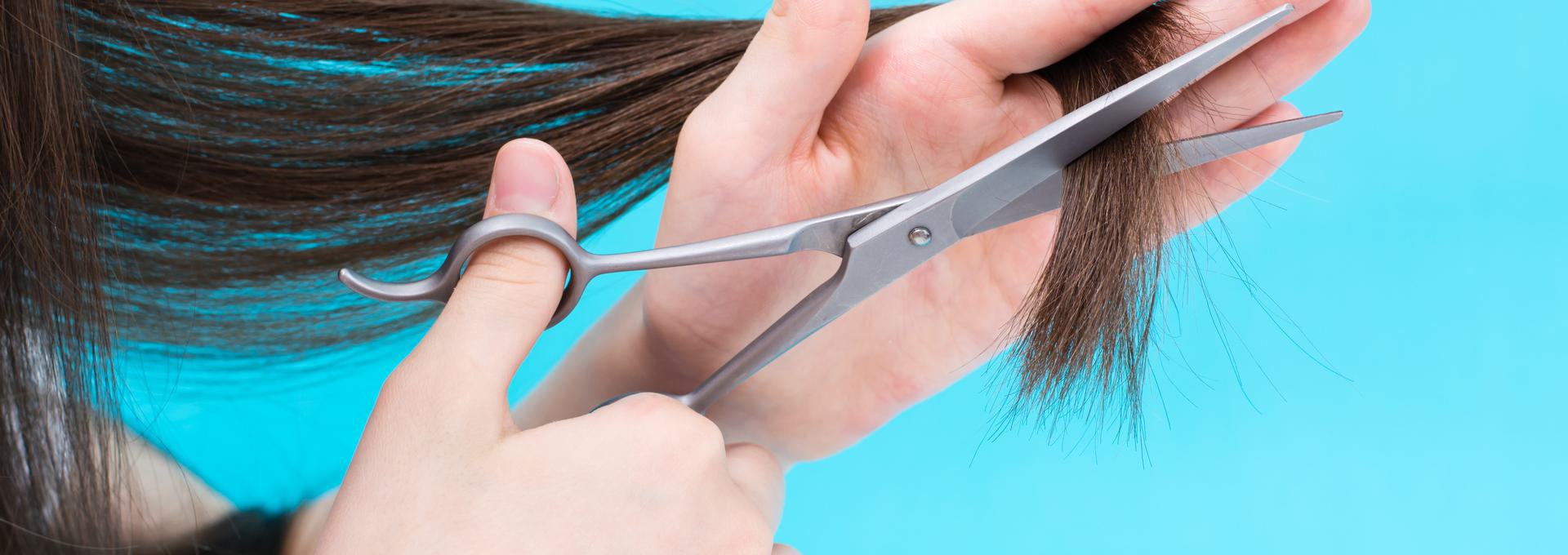 Close-up of hands cutting brown hair with scissors against a blue background.