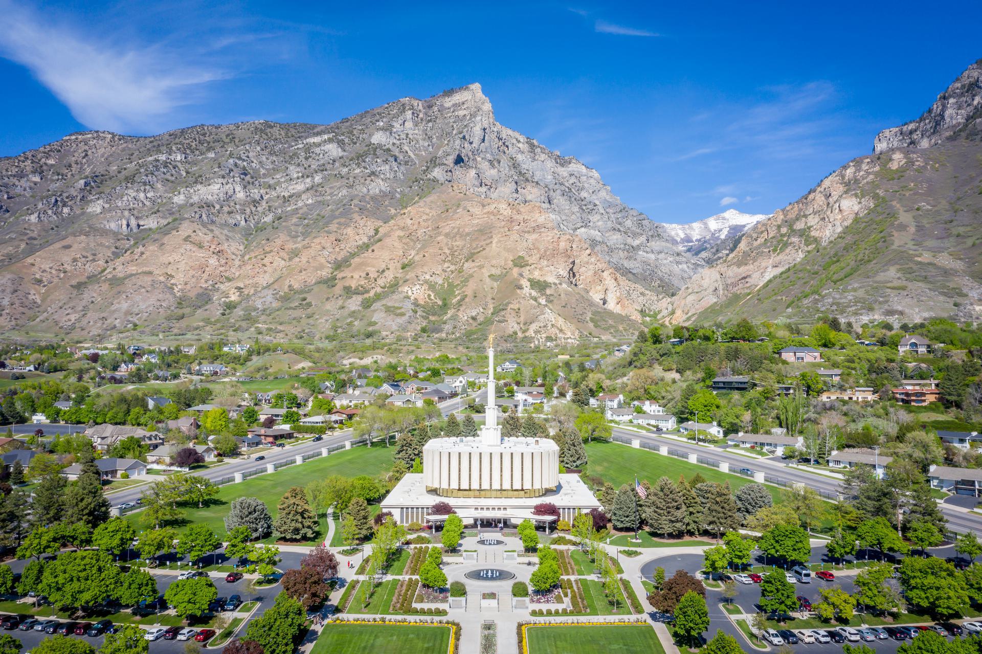 Temple and Mountain View Aerial view of a white temple with a spire surrounded by green lawns and trees, set against a backdrop of rugged mountains under a clear blue sky.
