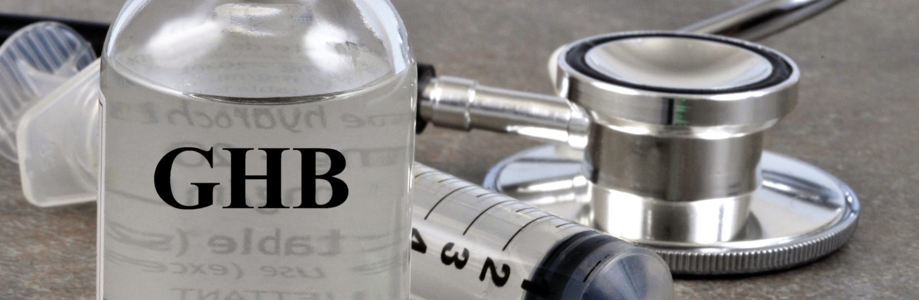 GHB Drug Test (Gamma-Hydroxybutyrate) Close-up of a bottle labeled GHB Drug Test (Gamma-Hydroxybutyrate) next to a syringe and stethoscope on a table.