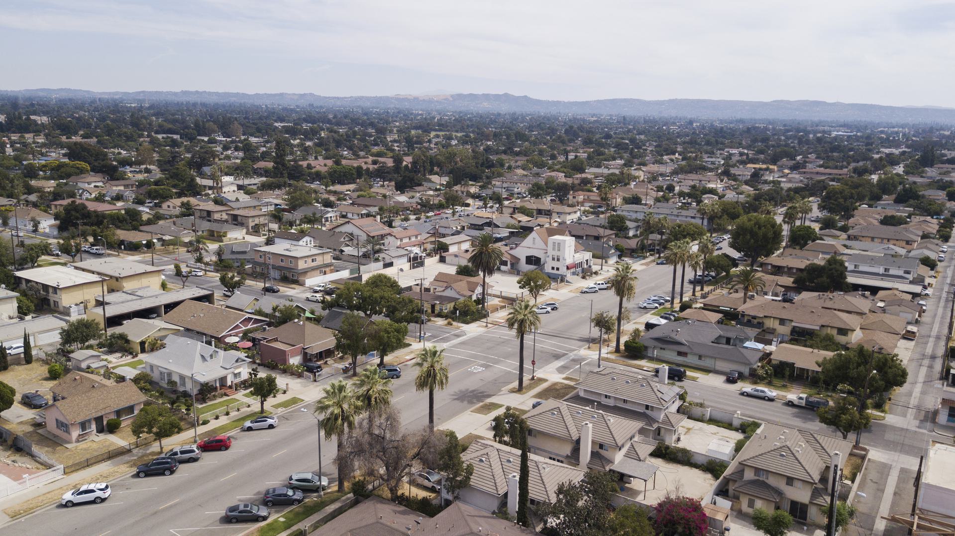 Suburban Neighborhood Aerial View Aerial view of a suburban neighborhood with rows of houses, streets lined with palm trees, and distant hills under a cloudy sky.