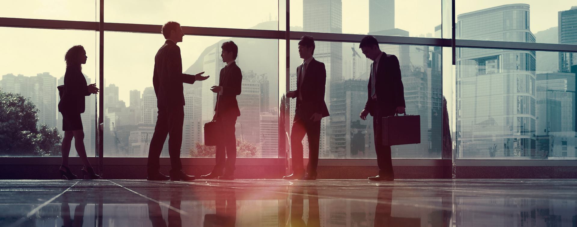 Silhouetted business people conversing in front of large windows with a city skyline backdrop.