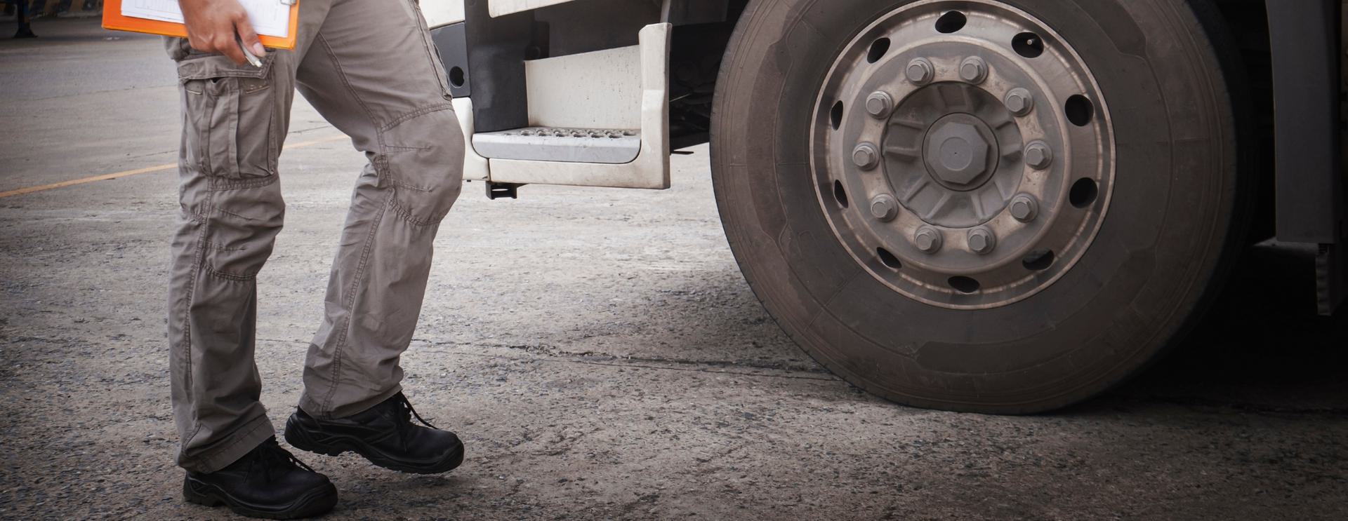 Person in cargo pants holding clipboard near truck tire on pavement.