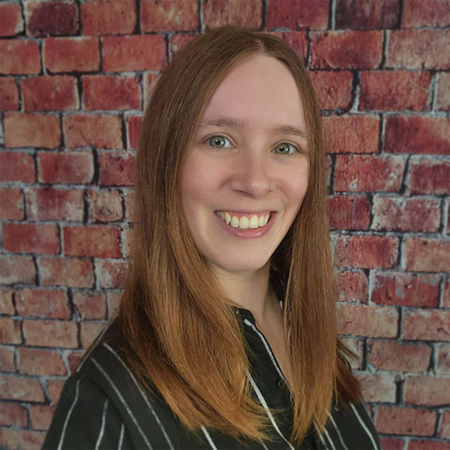 Nicole (Director of Digital Content and Marketing) Nicole smiling in front of a brick wall background, wearing a black and white striped shirt.