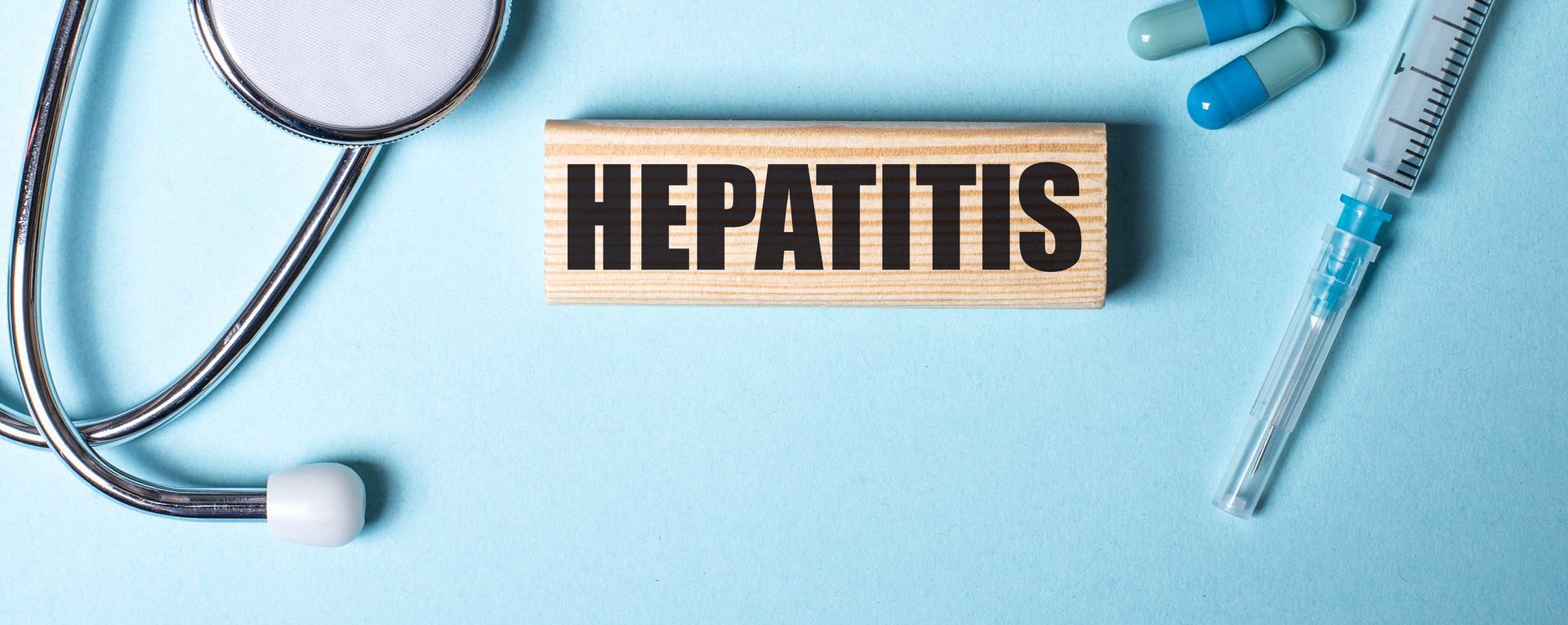 Stethoscope, pills, and syringe surrounding a wooden block with the word 'HEPATITIS' on a blue background.
