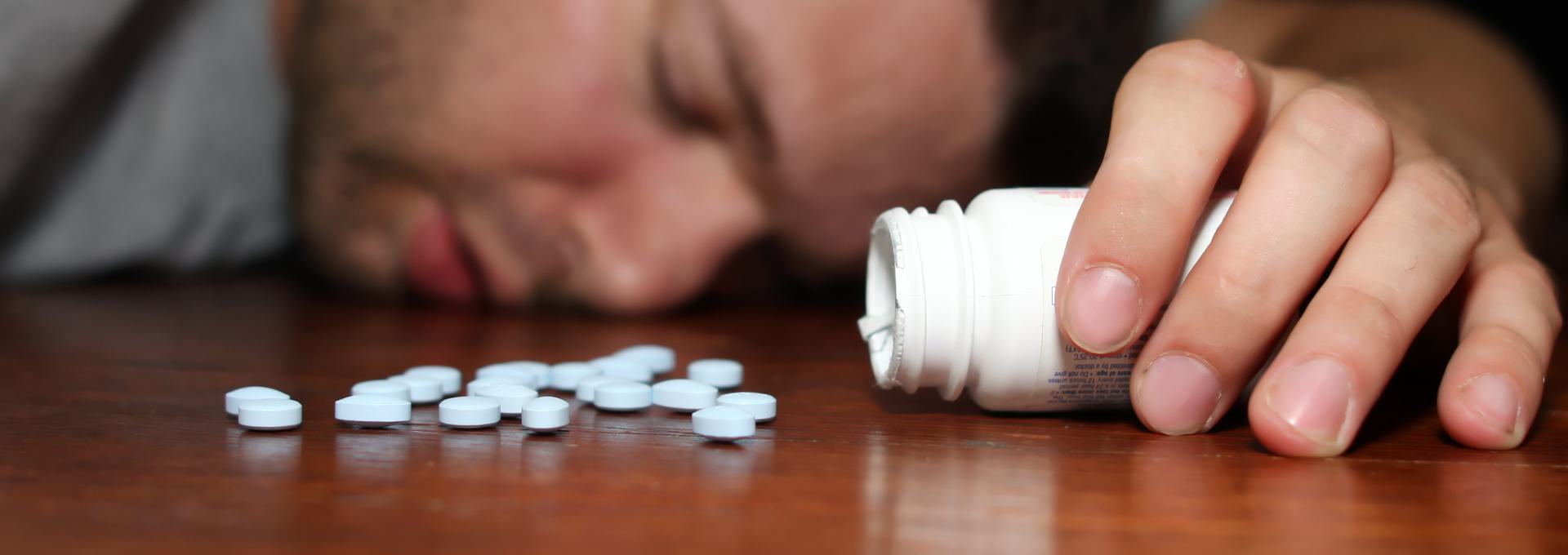 Ambien Drug Test (Zolpidem) Person lying unconscious next to spilled pills from a bottle on a wooden surface.