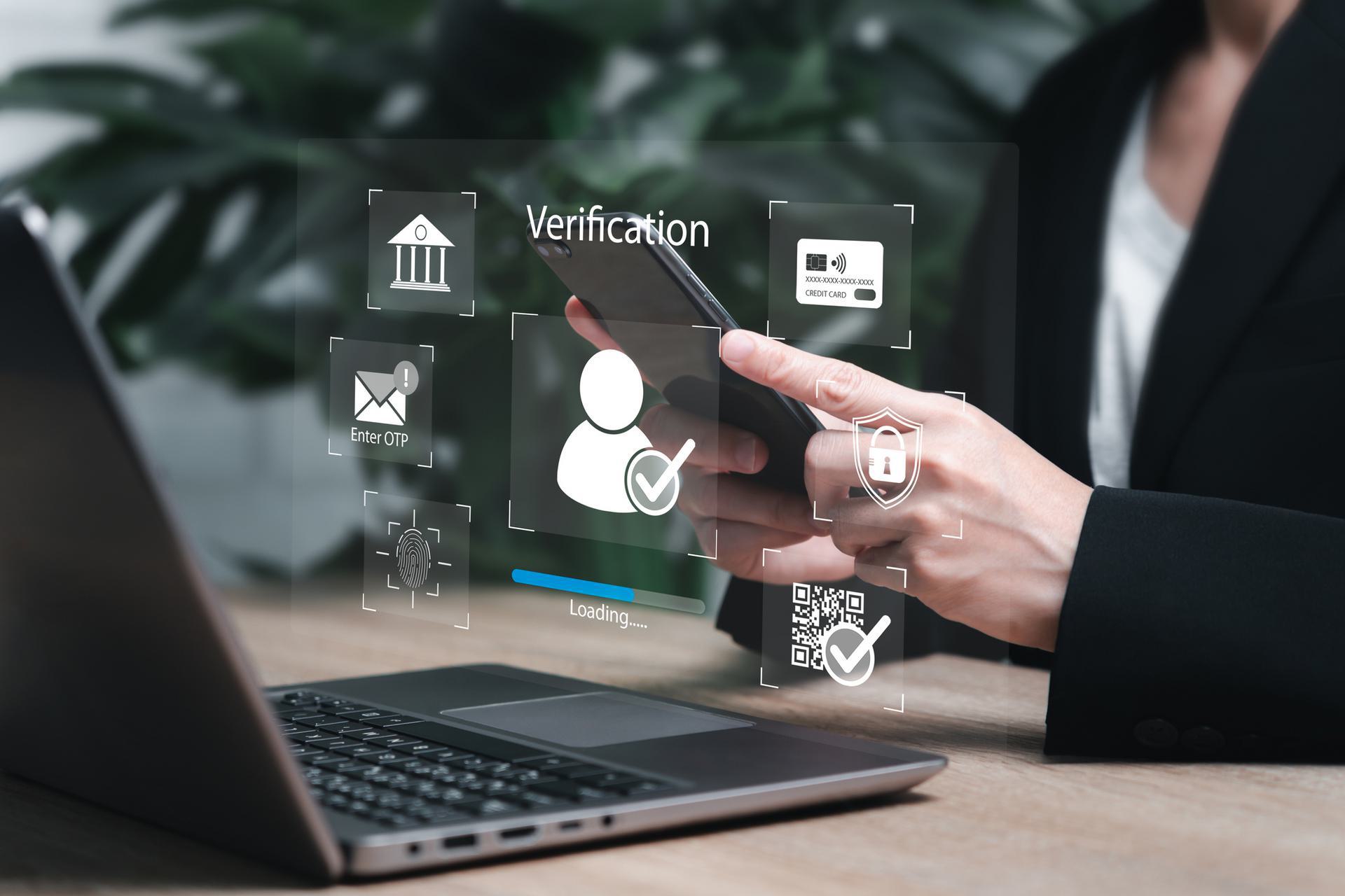 Person holding a smartphone with digital verification icons, including fingerprint and credit card, displayed above a laptop.