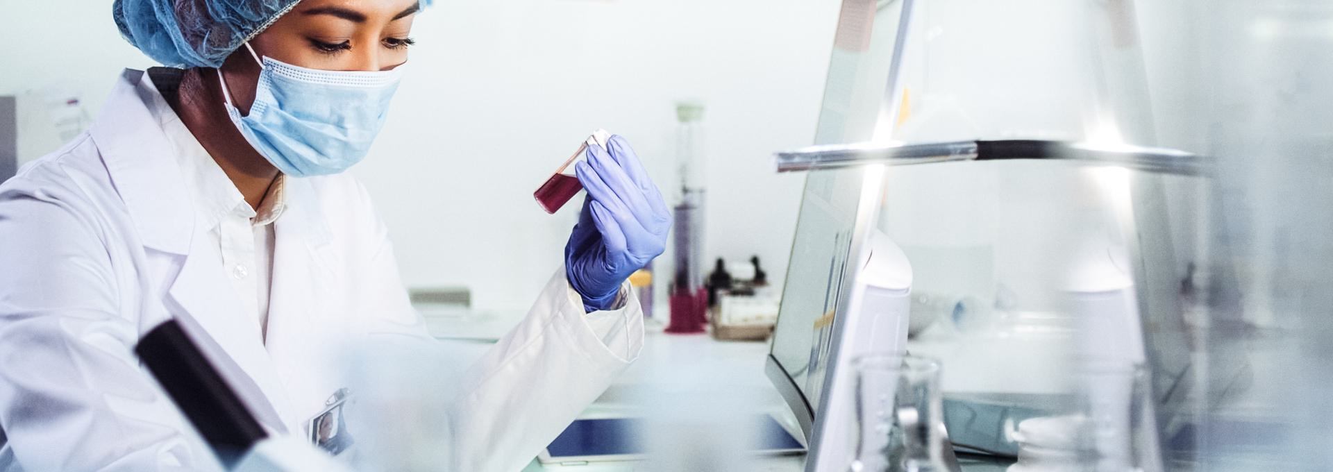Scientist in a lab coat and mask examining a vial of blood in a laboratory setting.