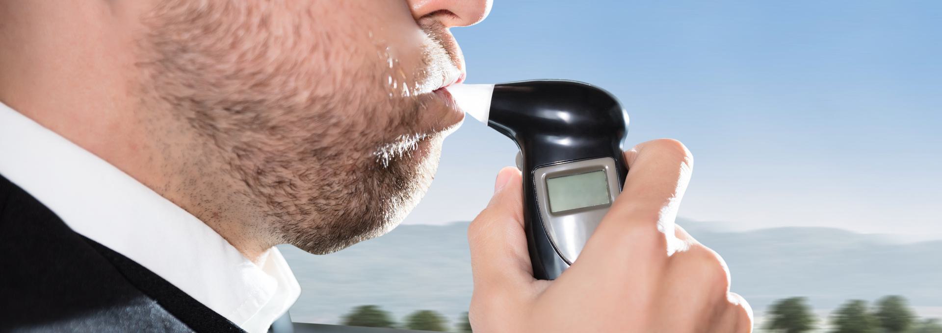 Person using a breathalyzer device outdoors with clear sky in the background.