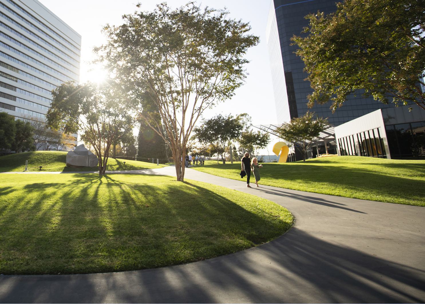 Sunlit urban park with trees, grass, and people walking on a path near modern buildings.
