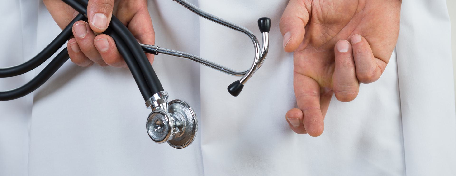 FACIS (Healthcare Fraud) Close-up of a doctor holding a stethoscope behind their back with crossed fingers, wearing a white coat.